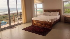 A bedroom with a bed, a wooden headboard, a nightstand, a red rug, and large windows offering a view of the ocean. - Modern Duplex 2-Bedroom Apartment with Sea View of Baroro Beach. (Bacnotan)