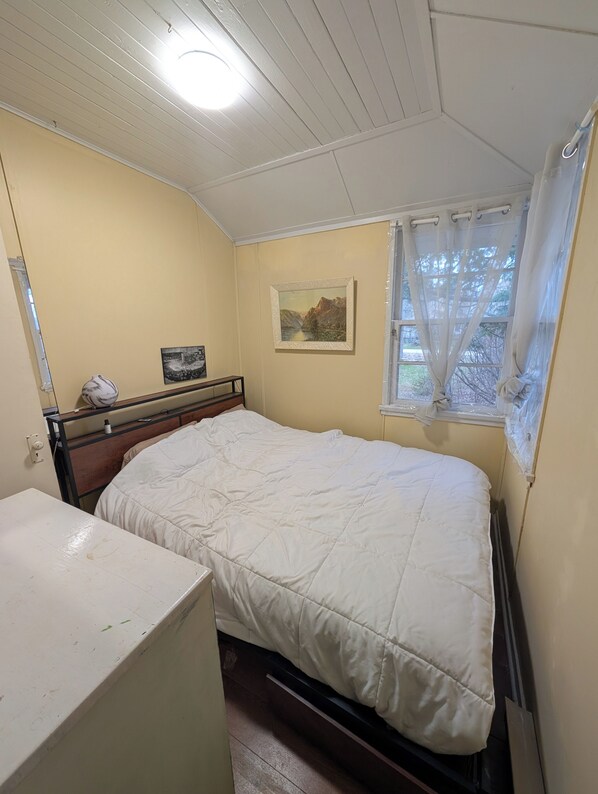 A bedroom with a bed, a bedside table, a window with curtains, a wall-mounted picture, and a ceiling light.