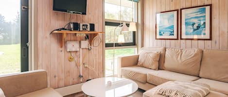 Una habitación acogedora con un sofá, una mesa redonda de vidrio, un televisor montado en la pared y cuadros enmarcados.