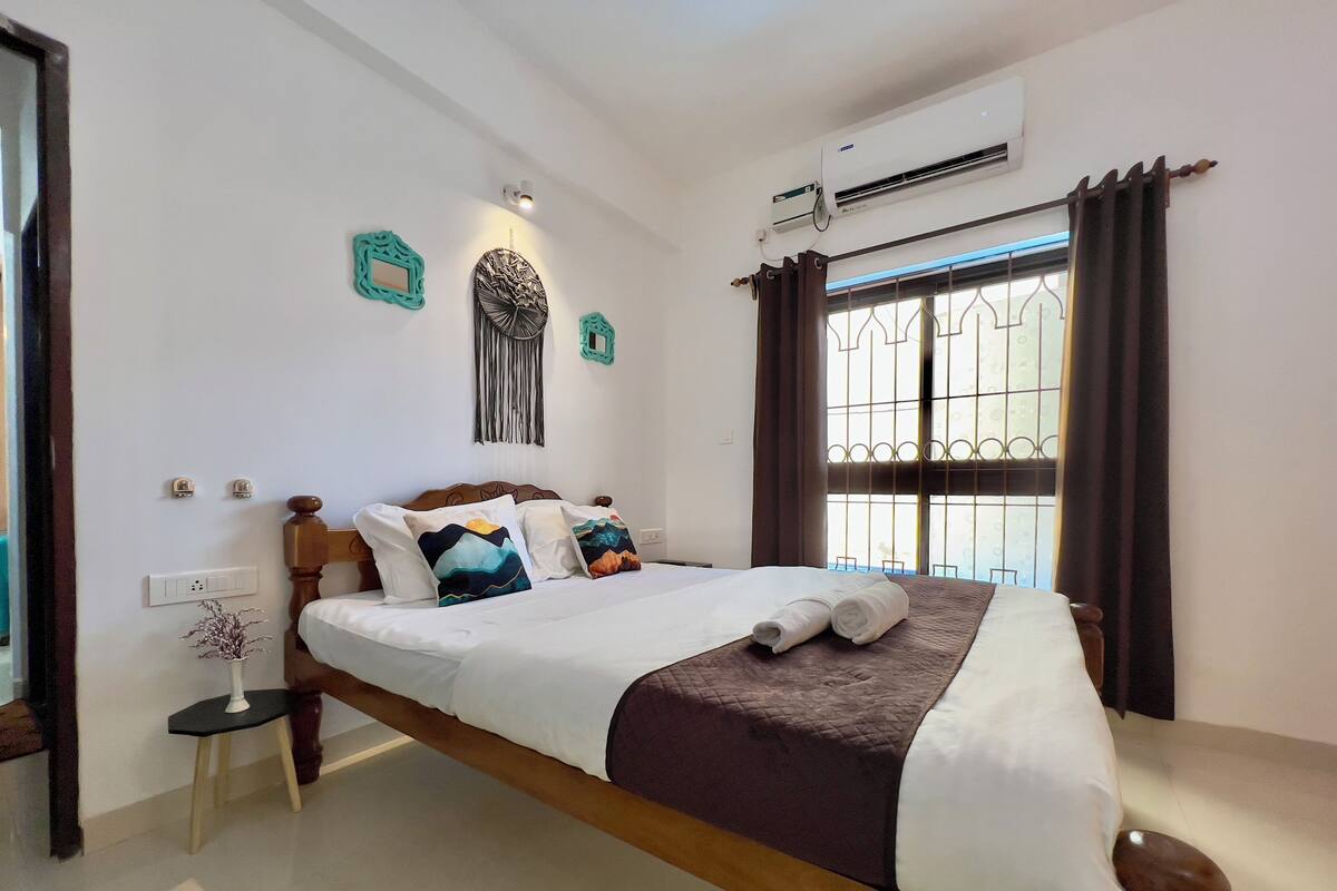 A neatly made bed with a brown headboard, white bedding, and decorative pillows. There is a small bedside table with a vase and a wall-mounted light. An air conditioning unit is visible on the wall, and a window with curtains is on the right side.
