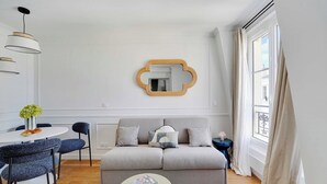 Un salon moderne avec un canapé gris, une table basse ronde, une table à manger avec des chaises et une grande fenêtre avec des rideaux.