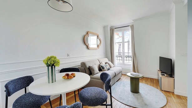 Un salon moderne avec une table à manger ronde, des chaises recouvertes de tissu bleu, un canapé beige, un meuble pour la télévision et une grande fenêtre avec des rideaux.