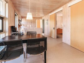 A modern interior with a wooden ceiling, a dining table with chairs, a sofa, and a kitchen area visible through an open door.