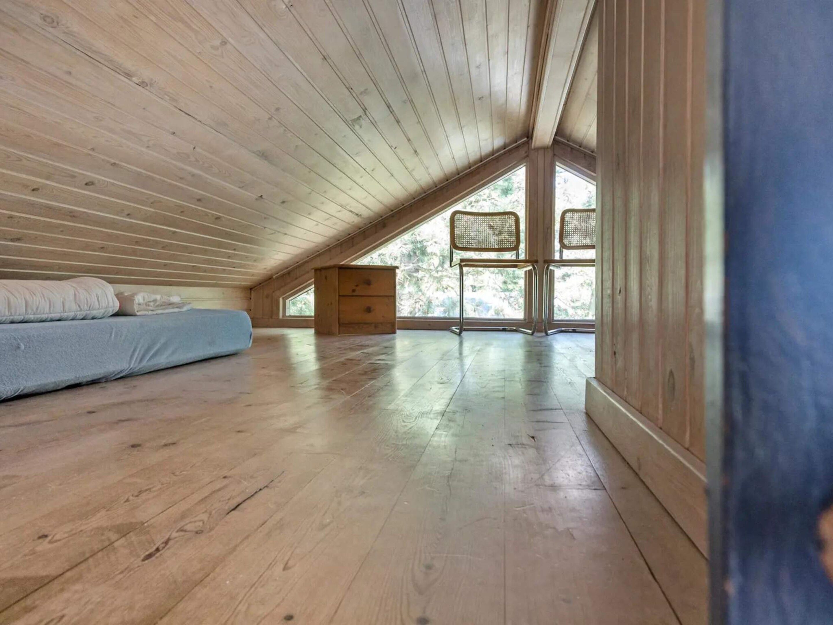 Amplia habitación en ático con pisos de madera, una cama elevadas y grandes ventanas que ofrecen vista a árboles.
