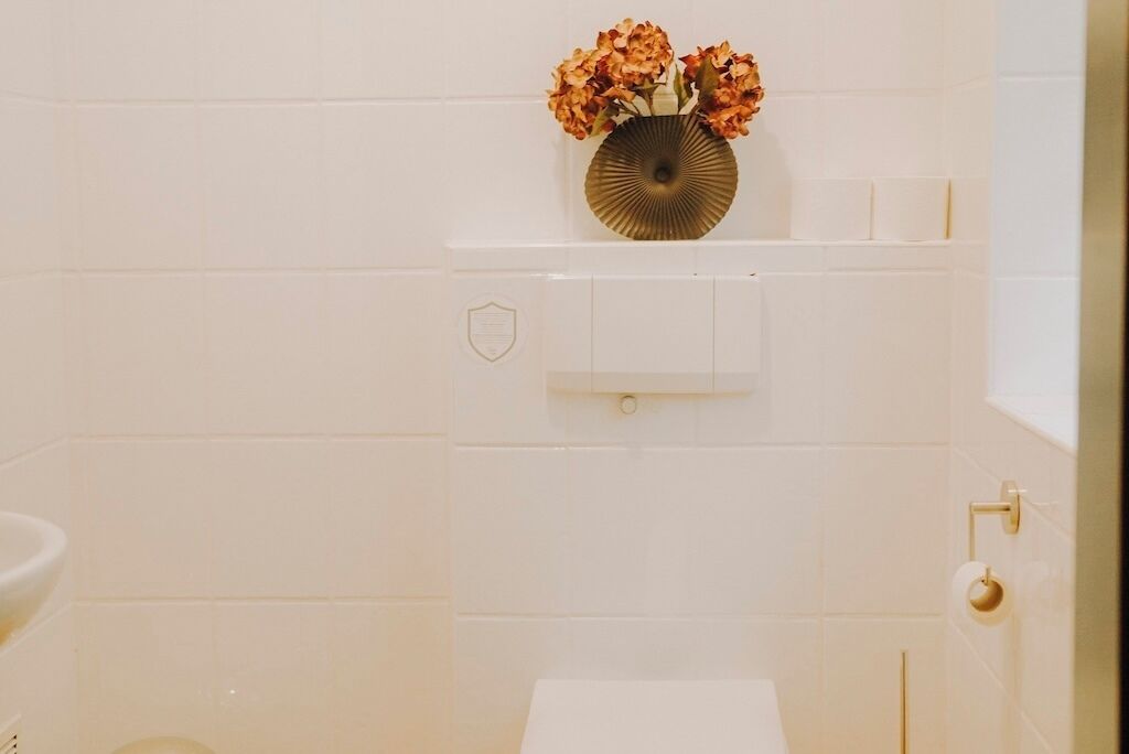 Et badeværelse med hvide fliser, toilet, vask og vægmonteret toiletpapirholder.