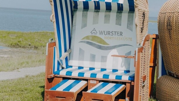 En strandstol med en stribet pude og et logo, placeret på en græsplæne med udsigt til havet.