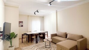 A living room with a beige sofa, a dining table with chairs, a small side table, a potted plant, and a patterned rug.