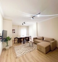 A living room with a beige sofa, a dining table with chairs, a small side table, a potted plant, and a patterned rug.