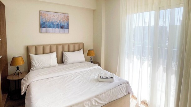 A hotel room with a large bed, two bedside tables with lamps, a painting on the wall, and sheer curtains on the window.
