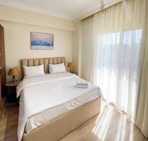 A hotel room with a large bed, two bedside tables with lamps, a painting on the wall, and sheer curtains on the window. - Settle Inn Rawda Apartments (New Cairo)