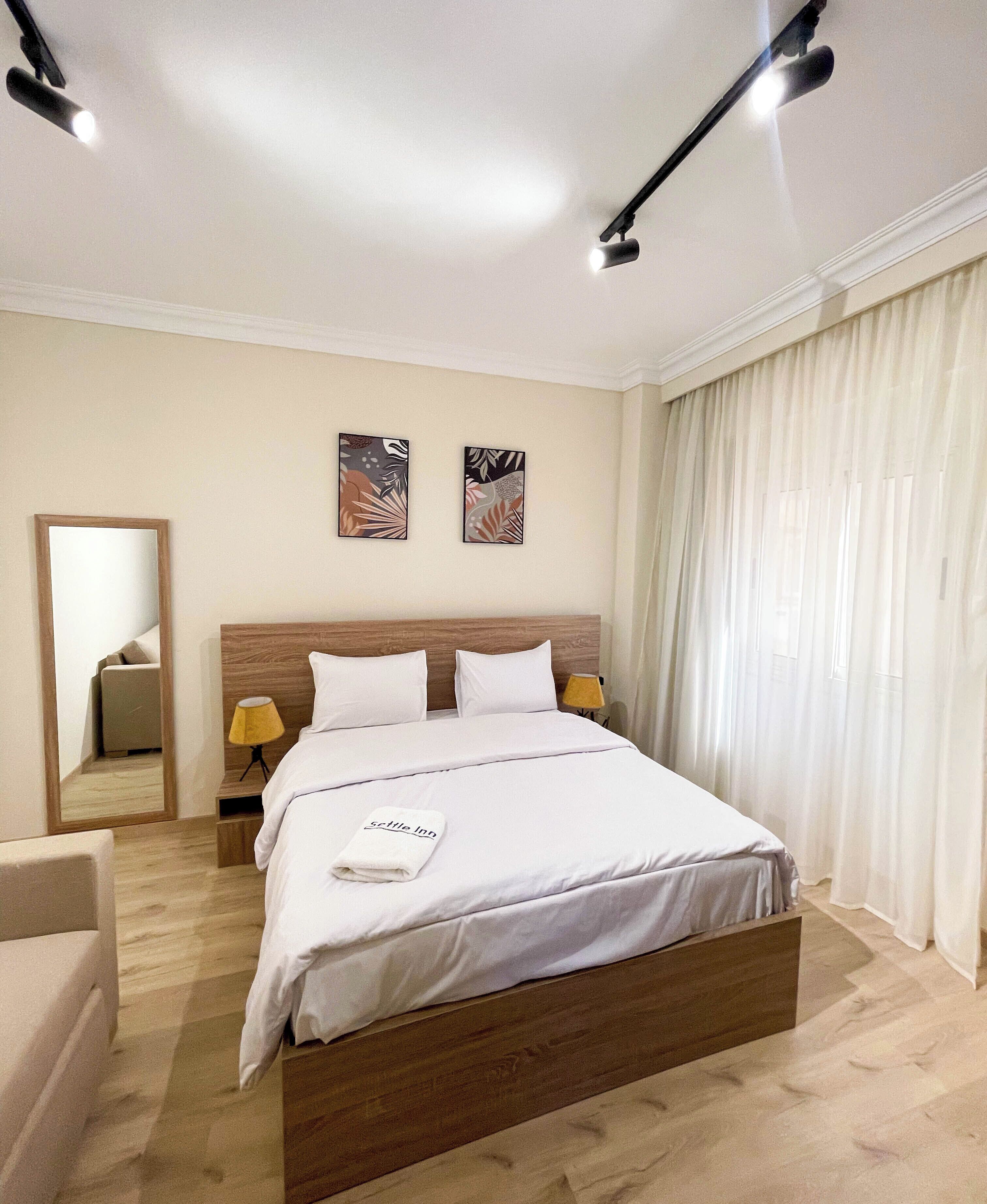 A modern hotel room with a large bed, two wall-mounted lamps, a mirror, and abstract artwork.