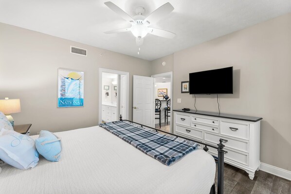 Une chambre à coucher comprenant un lit, un ventilateur de plafond, un téléviseur, une commode et une porte donnant sur une autre pièce.
