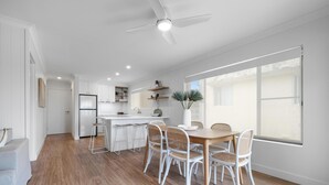 A modern kitchen with white cabinets, a built-in fridge, and a dining area with a wooden table and wicker chairs. - Sundaze at Kingy Creek (Kingscliff)