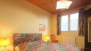 Un dormitorio con colcha estampada, cabecera de madera, mesita de noche con lámpara y ventana con cortinas.