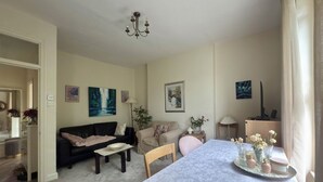 A living room with a dining table, a black sofa, a beige armchair, and a coffee table. There are paintings on the walls and a chandelier above the table.
