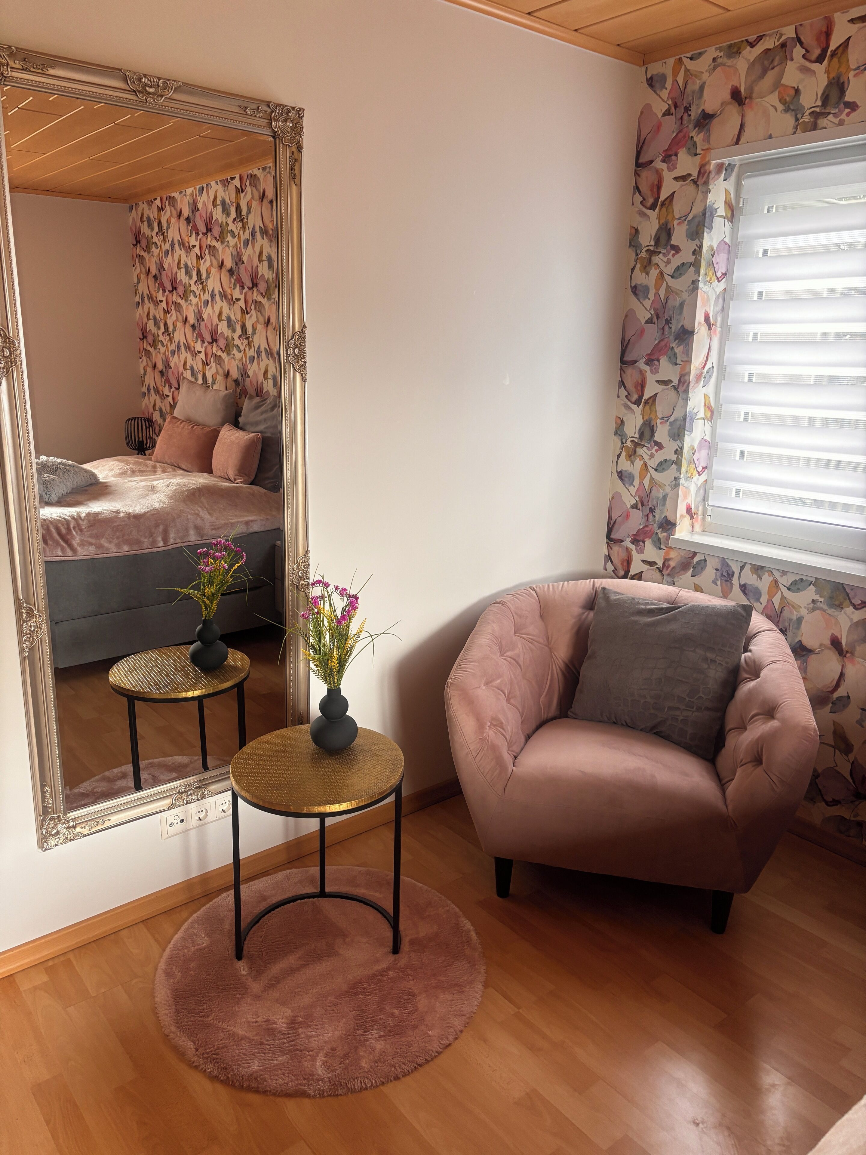 Un dormitorio con un espejo grande, una cama con papel tapiz floral, una mesa redonda con un florero, un sillón rosa y una ventana con persianas.
