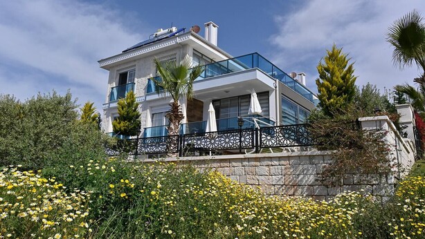 Ein modernes, zweistöckiges Hotel mit Balkon, umgeben von üppiger Vegetation und gelben Blumen.