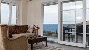 Ein Wohnzimmer mit einer Couch, einem Couchtisch und einer Blumenvase. Ein großes Schiebeglasfenster führt auf einen Balkon mit Blick auf das Meer.