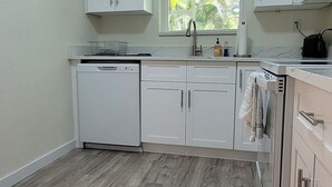 Una cocina con armarios blancos, lavavajillas, ventana con persianas y suelo de madera.