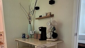 Una habitación de esquina con piso de madera, un mueble blanco, una estantería con utensilios de cocina y un florero con ramas.