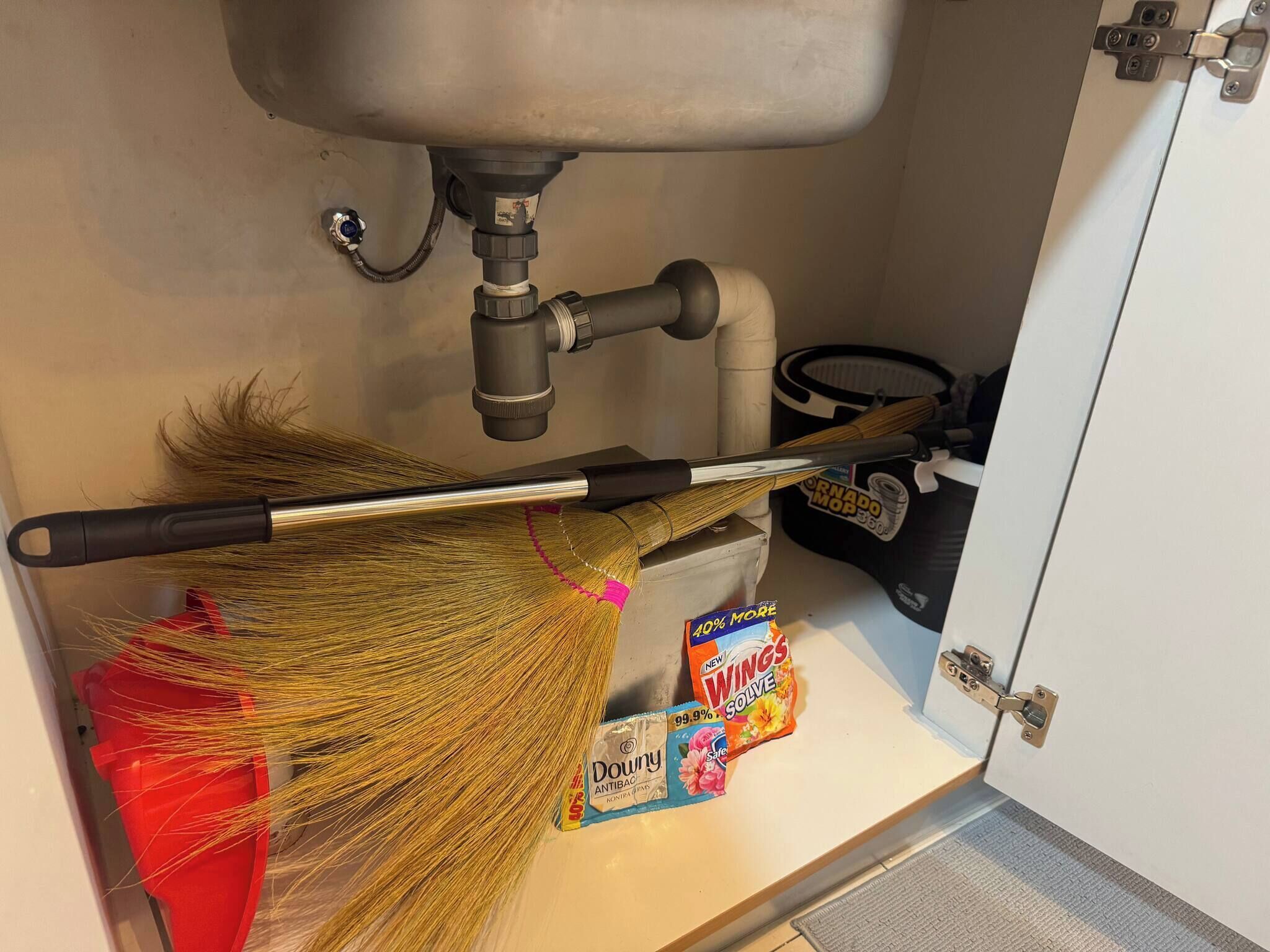 Un balai appuyé contre un évier, une serpillière et une boîte de produit nettoyant Windex sous un meuble.