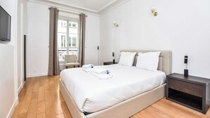 Ein Hotelzimmer mit einem großen Bett, einem Nachttisch, einem an der Wand montierten Fernseher und einem Fenster mit Vorhängen.