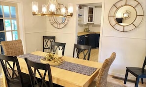 Un coin repas comprenant une table en bois, des chaises noires, une chaise en osier, un lustre et une horloge murale.