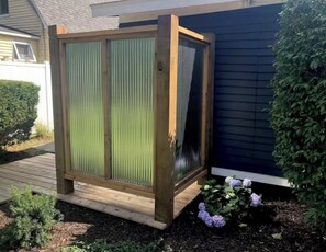 Une cabine de douche extérieure en bois, dotée d’une porte en métal, située à côté d’une maison dont le revêtement extérieur est de couleur sombre.