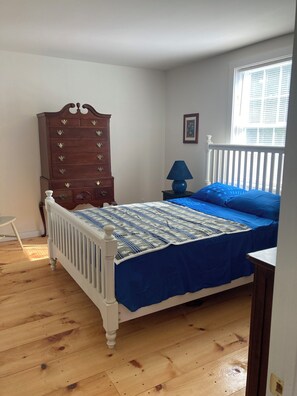 Ein Schlafzimmer mit einem weißen Bett, blauer Bettwäsche, einem hölzernen Schminktisch, einem Nachttisch mit Lampe und einem Fenster mit Jalousien.