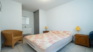 A bedroom with a bed, a chair, a bedside table, a lamp, and a bathroom visible through an open door. - Apartment in Valras Plage With View (Sérignan)