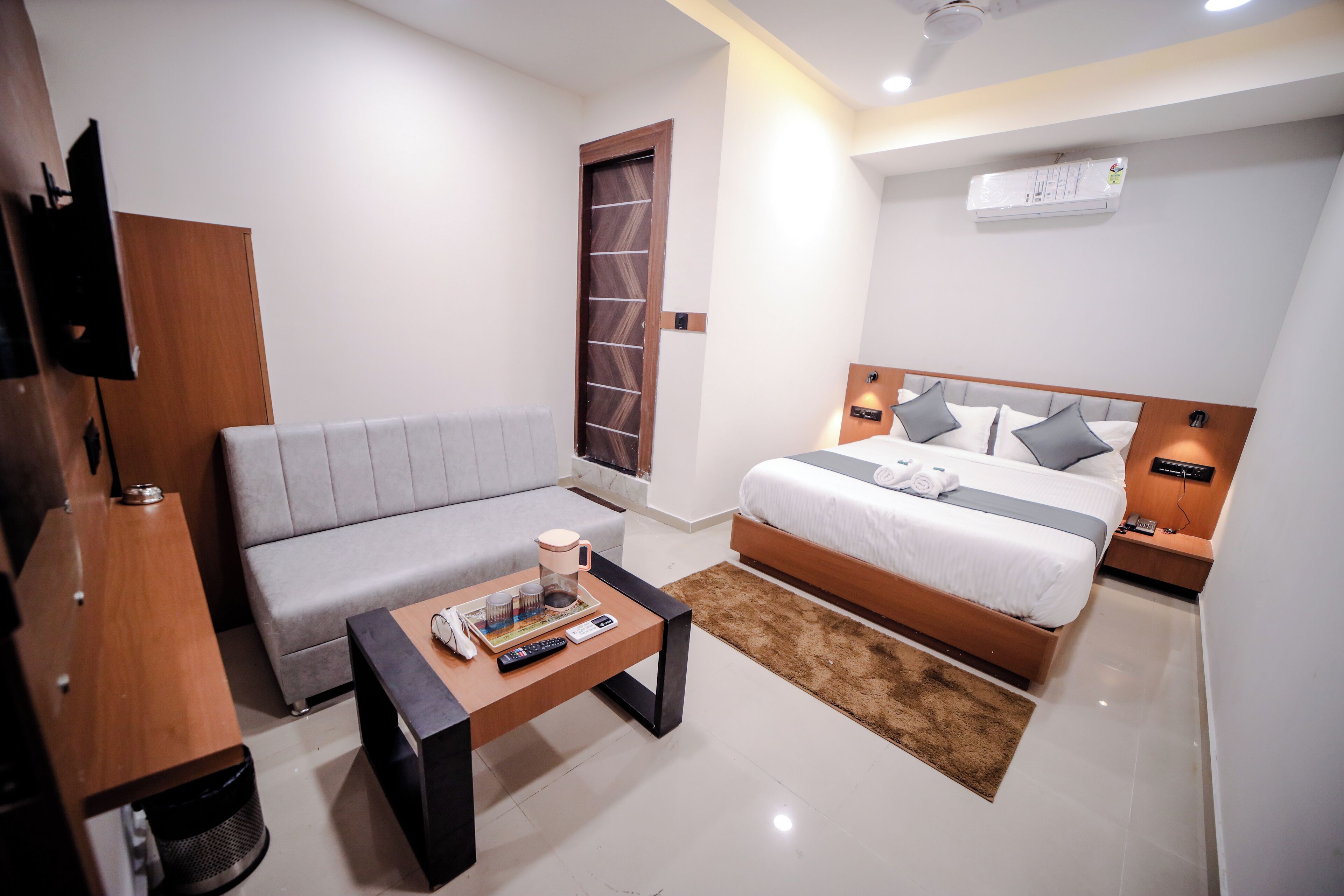 Una habitación de hotel moderna con sofá, una cama con cabecera de madera, una mesa de centro y un televisor montado en la pared.