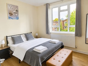 A bedroom with a bed, bedside tables, a bench, a window with curtains, and a view of buildings outside. - Cozy 3 BDR flat in London, tube, free parking, perfect for workers and tourists (London)