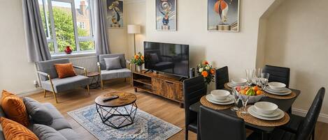A modern living room with a sofa, armchair, coffee table, TV, and dining area with chairs and table set for a meal.
