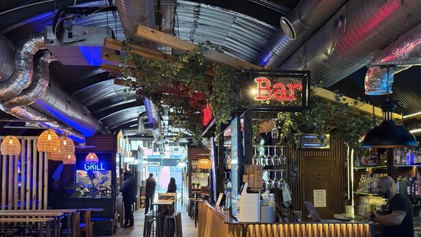 Un bar intérieur avec un panneau lumineux au néon, un comptoir en bois et des luminaires suspendus.
