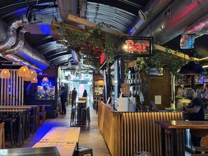 Un bar intérieur avec un panneau lumineux au néon, un comptoir en bois et des luminaires suspendus.