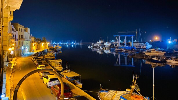 A nighttime marina with boats docked, a road with cars, and buildings along the waterfront.