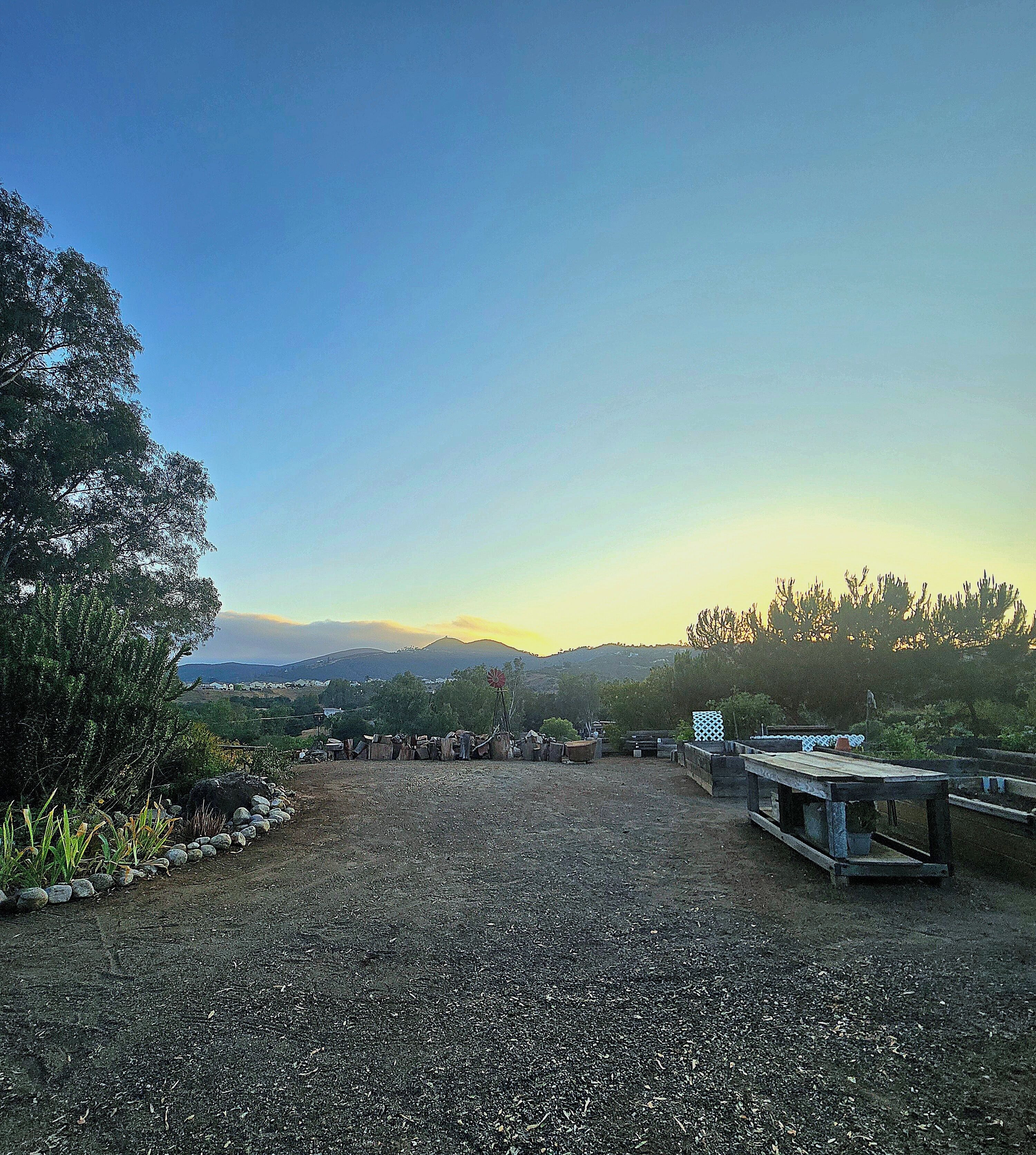 A gravel area with wooden benches and a windmill, surrounded by trees and a distant mountain range.