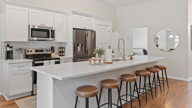 Une cuisine moderne avec des meubles blancs, des appareils en acier inoxydable, un îlot central doté de tabourets et un grand miroir rond accroché au mur.