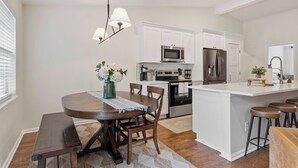Une cuisine moderne avec des meubles blancs, un îlot central doté de tabourets, une table à manger avec des chaises et un luminaire suspendu.