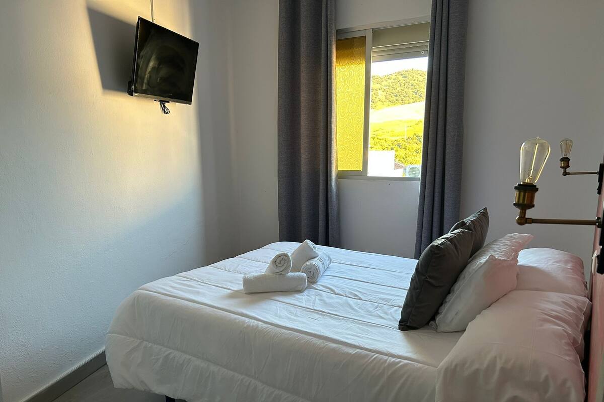 A neatly made bed with white linens and a rolled towel, a bedside table with a lamp, and a window with curtains.