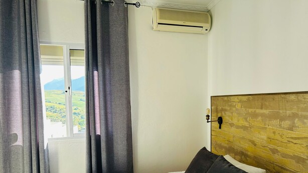 A hotel room with a bed, a window with curtains, an air conditioning unit, and a view of the outdoors. - Hostal El pinar (Montecorto)