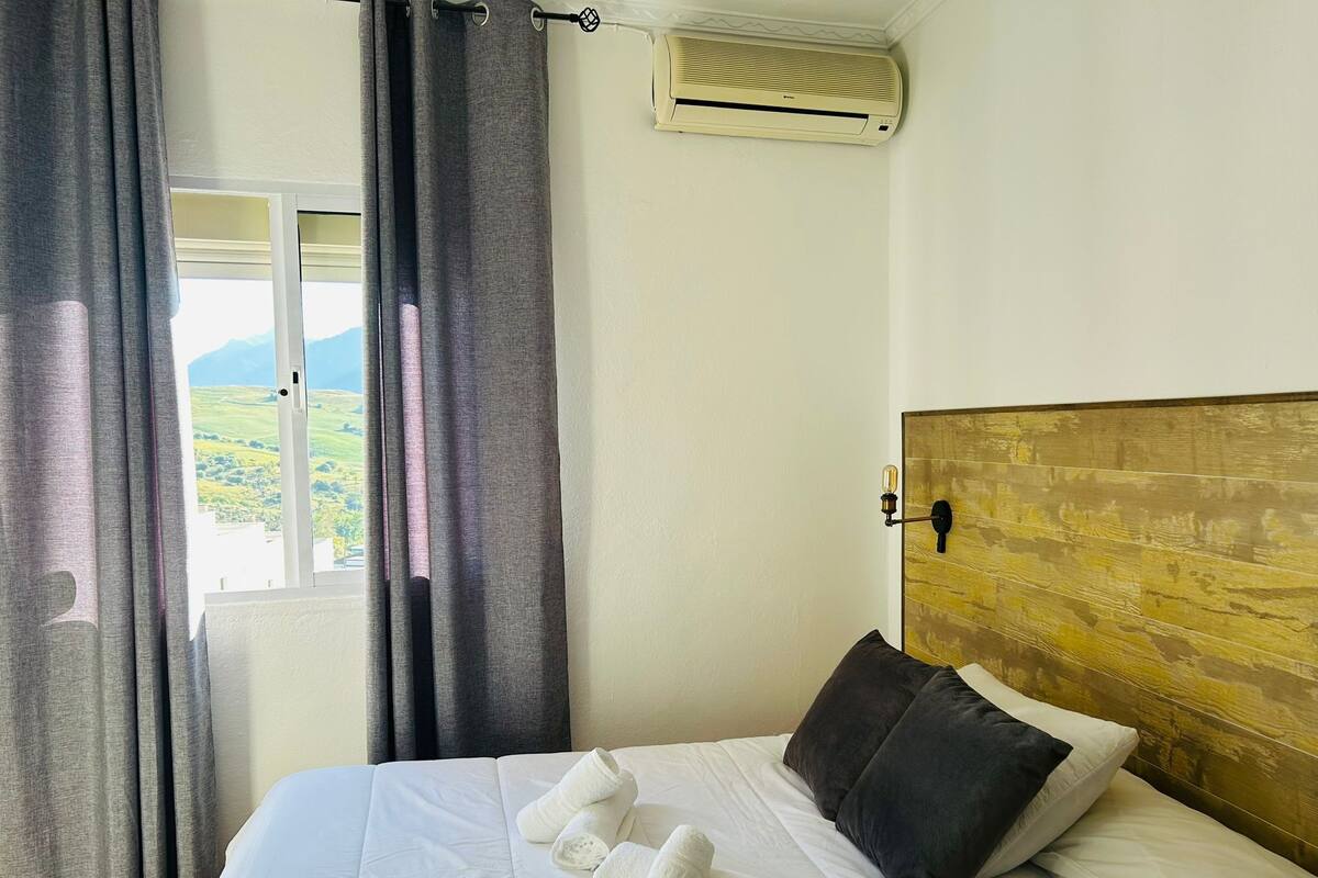 A hotel room with a bed, a window with curtains, an air conditioning unit, and a view of the outdoors.