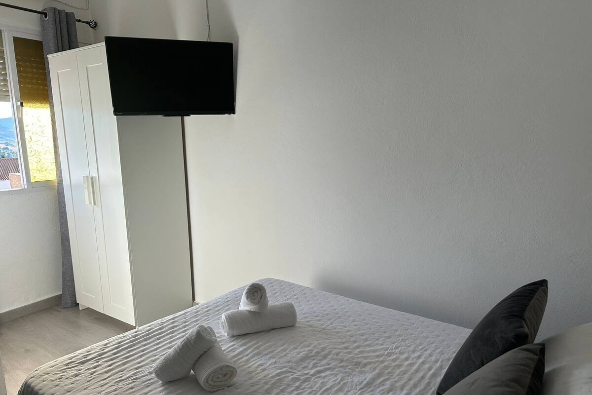 A neatly made bed with rolled towels, a flat-screen TV mounted on the wall, and a white wardrobe.