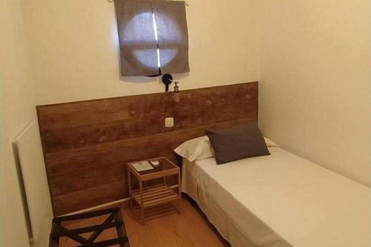 A small, single-bed room with a wooden headboard, a bedside table, a nightstand, and a wall-mounted light fixture.