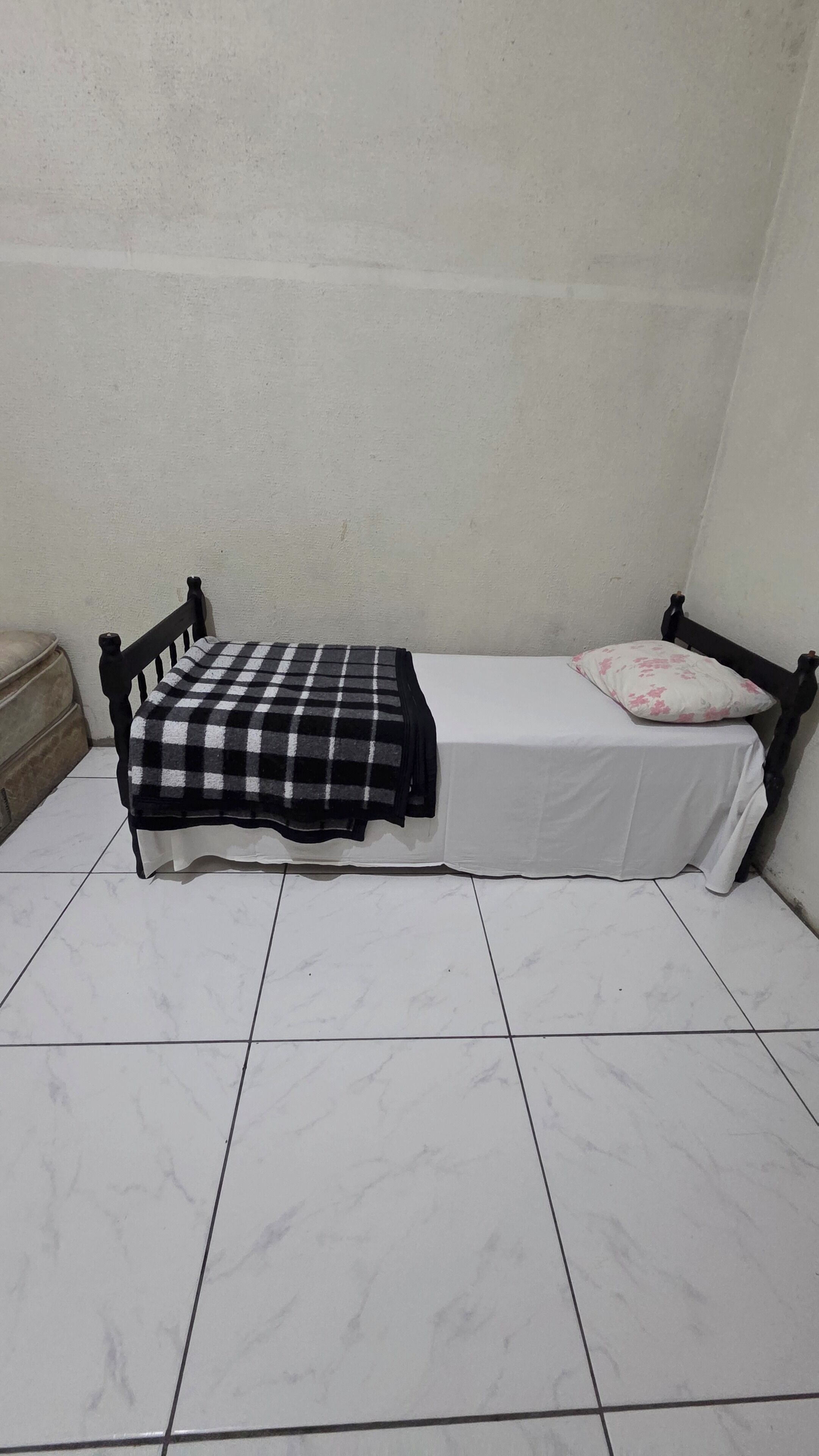 Uma cama pequena com um cobertor xadrez e um único travesseiro, apoiada em uma parede lisa com piso de azulejos.