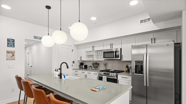 Une cuisine moderne dotée d’un îlot central, de tabourets de bar, d’appareils électroménagers en acier inoxydable et de luminaires suspendus.