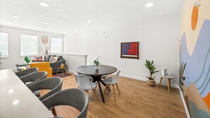 Ein modernes, gut beleuchtetes Zimmer mit Essbereich, der einen runden Tisch und graue Stühle, eine Couch und ein farbenfrohes Wandbild umfasst.