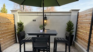 Eine Terrasse im Freien mit Tisch und vier Stühlen, einem kleinen Topfpflanzen und einer an der Wand befestigten Leuchte.