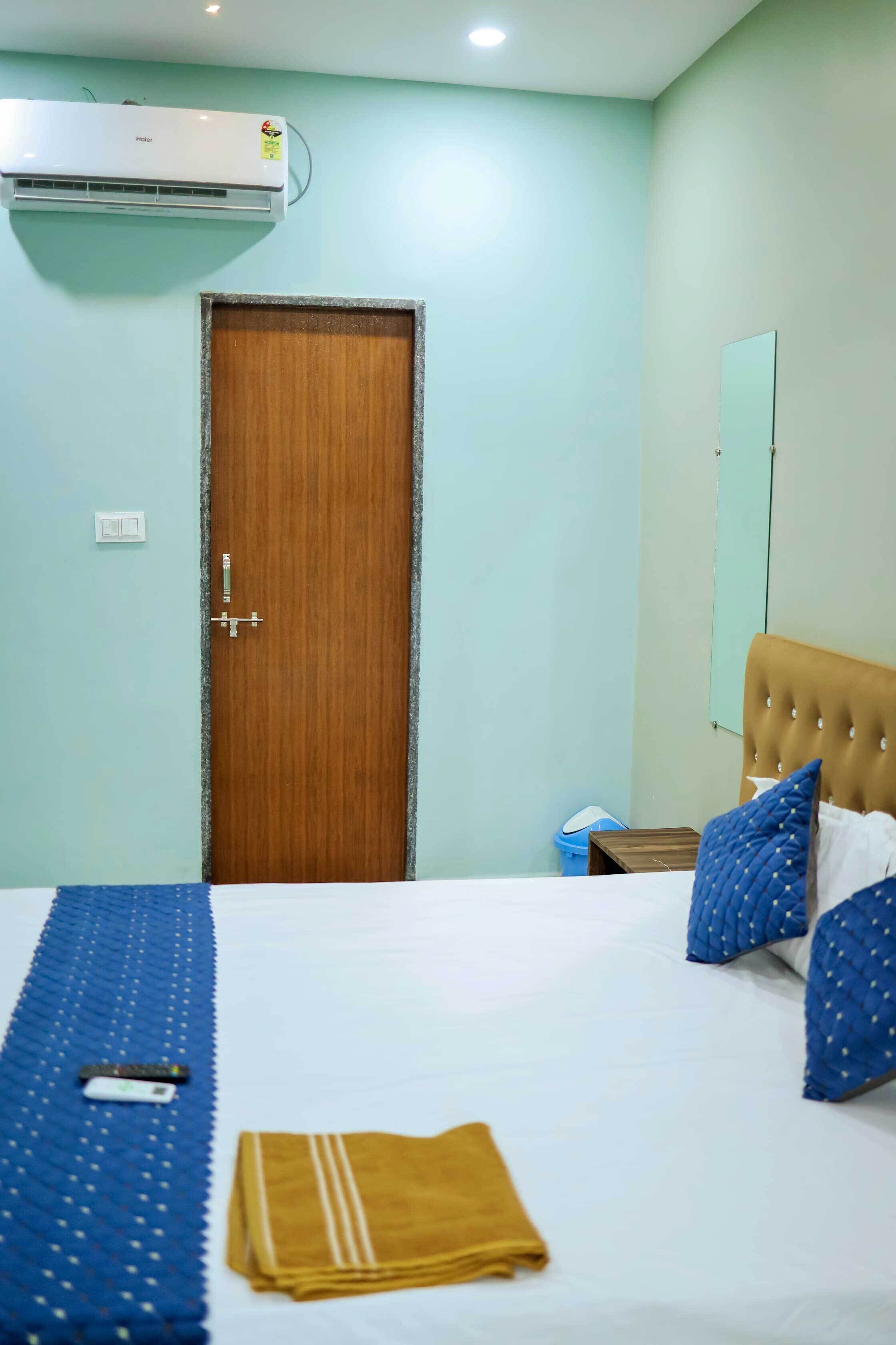 A hotel room with a bed, a wooden door, a wall-mounted air conditioner, a bedside table, and a mirror.