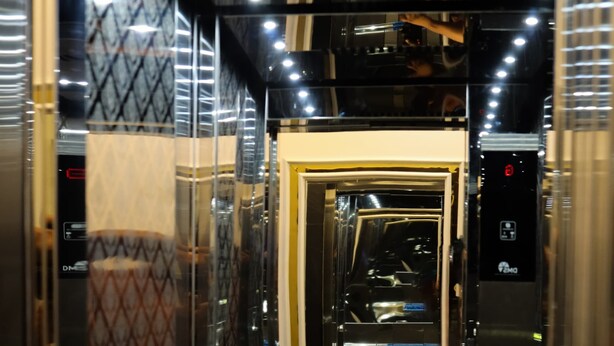 An interior view of an elevator with a metallic finish, control panel, and a door leading to another area. - Hotel Shivganga (Ujjain)